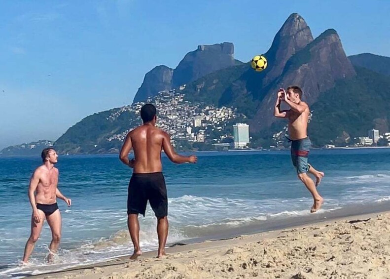 Three people playing soccer on a beach with a mountain in the background. 1