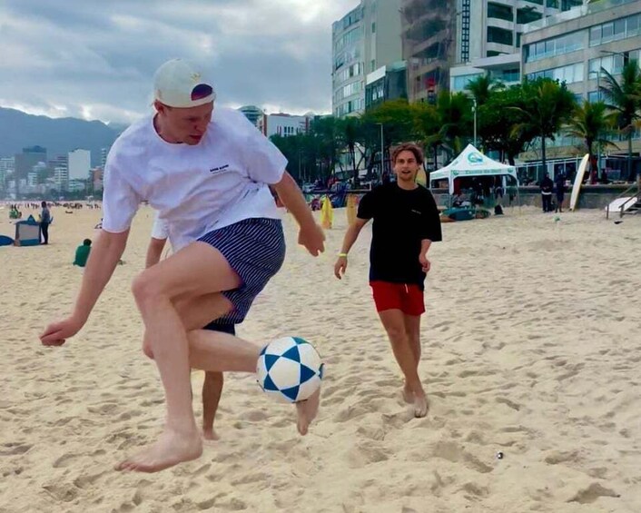 A person kicking a soccer ball on a sandy beach with palm trees and buildings in the background. 5