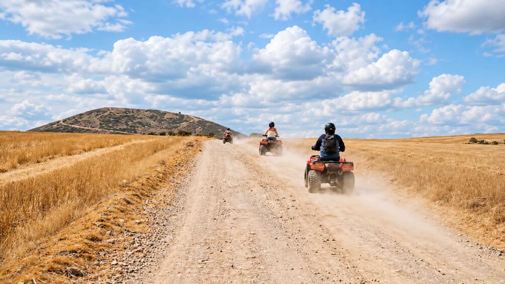 Three people riding ATVs on a dirt road in a dry, open landscape with sparse vegetation and a hill in the background. 5