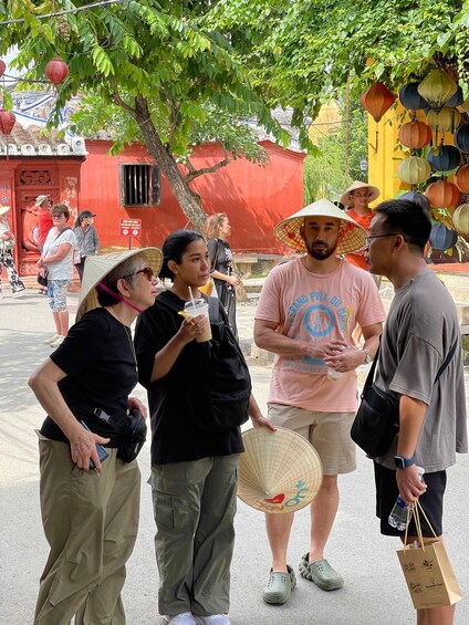 A group of people standing outdoors near a red building with traditional architecture, holding drinks and a fan. 3