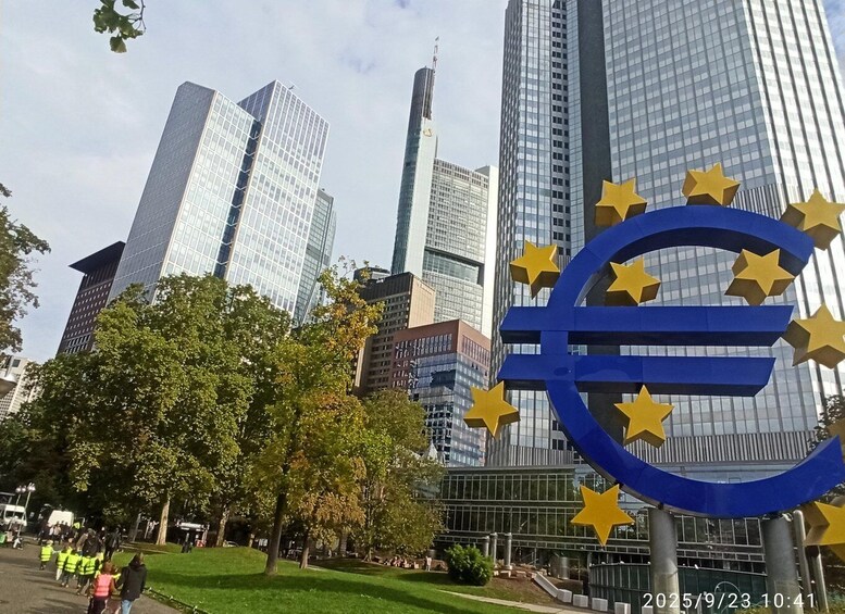 A cityscape with modern skyscrapers and a prominent Euro symbol sculpture. 1