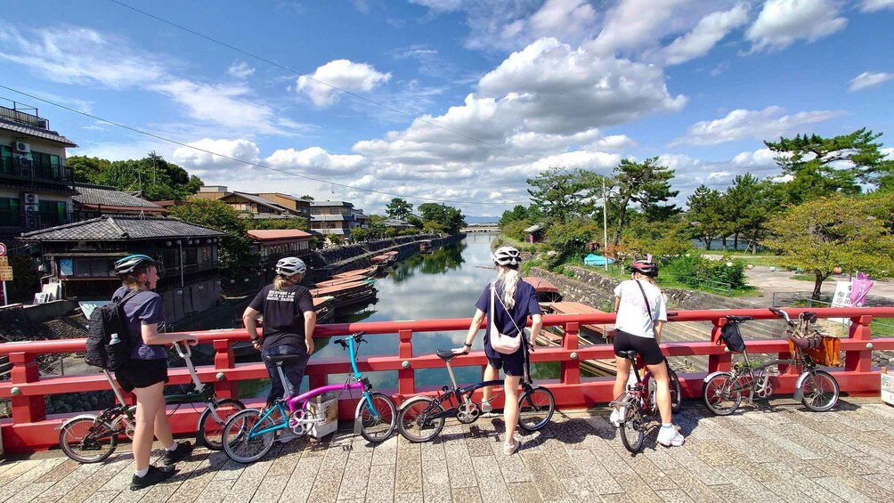 A group of cyclists on a bridge with a river and traditional buildings in the background. 1