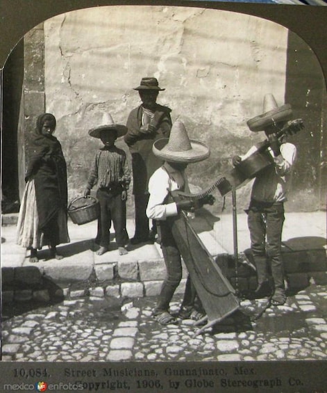 A street musician, a woman with a basket, and a man with a large hat standing on a cobblestone street. 3