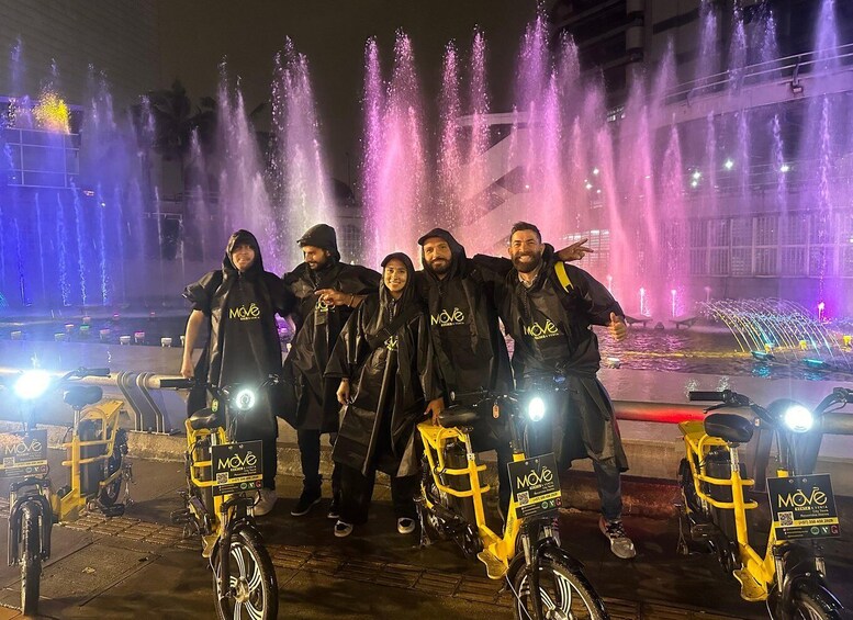 A group of people on bicycles with 'MOVE' branding, standing in front of a fountain with colorful lights at night. 1