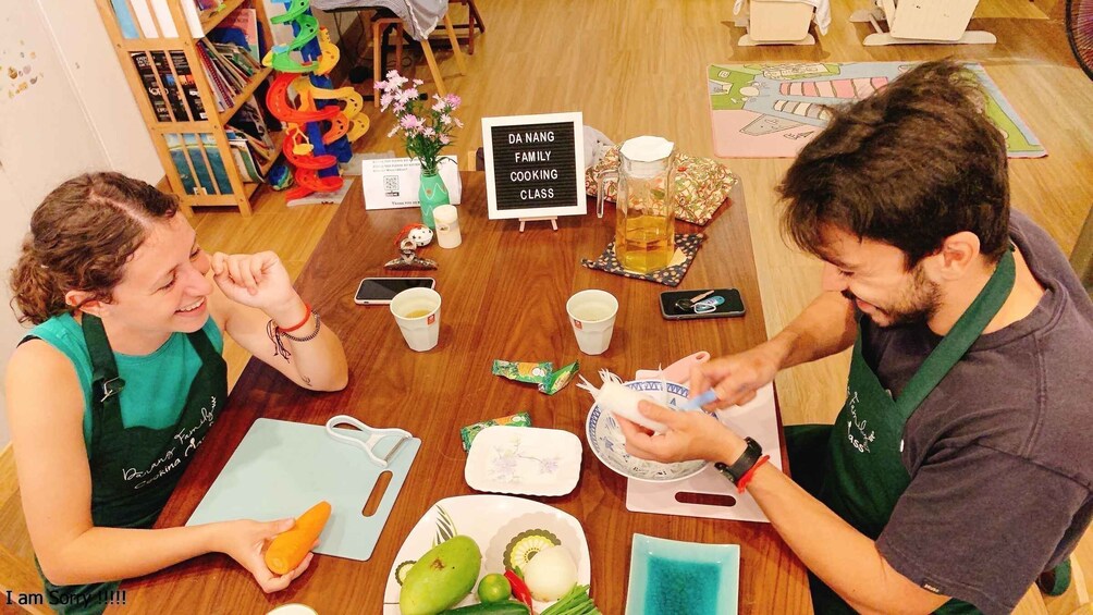 Kaksi ihmistä istuu pöydän ääressä, pöydällä on ruokaa ja juomia. Taustalla näkyy kyltti, jossa lukee ”DA NANG FAMILY COOKING CLASS”, ja värikäs lelu. 5