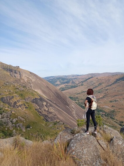 A person standing on a rocky outcrop overlooking a valley with hills and sparse vegetation. 1