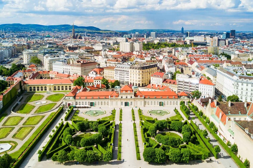 A cityscape with a prominent historical building surrounded by well-manicured gardens and a large cathedral in the background. 2