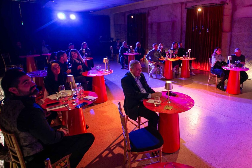 A formal indoor event with round tables and chairs, people seated and standing, and a stage in the background. 5
