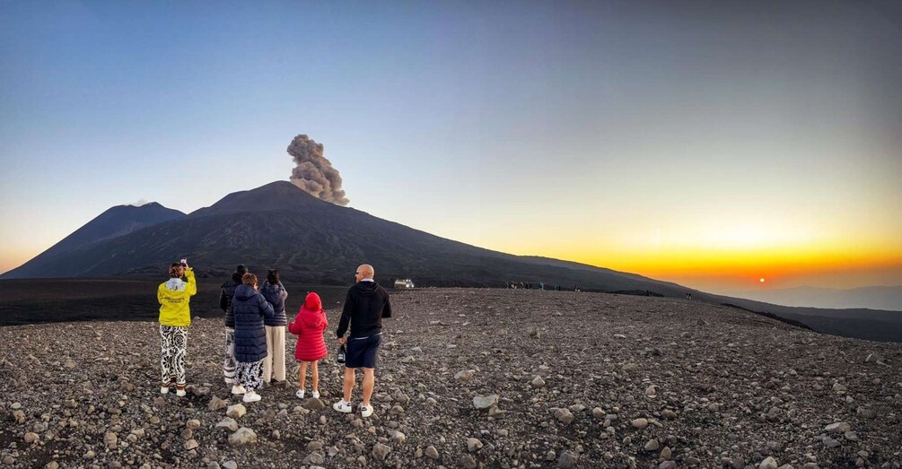 A group of people observing a volcano with smoke at its peak during sunset. 2