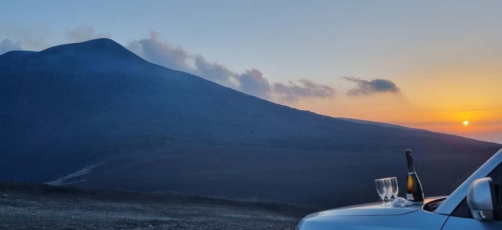 A car with a bottle and glass on the dashboard, a mountain in the background, and a sunset. 3