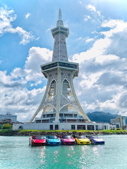 A tower with intricate lattice work and a modern building at its base, surrounded by a body of water with colorful cars parked in front. 4