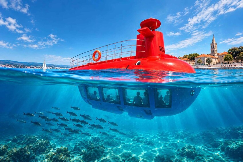 A red submarine-like vessel with passengers, submerged in clear blue water with a coastal town and a sailboat in the background. 1