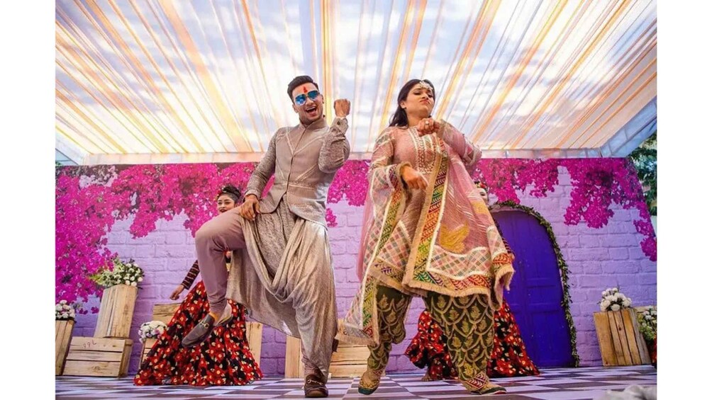 A couple in traditional attire dancing in a decorated indoor setting with a purple brick wall and pink flowers. 5