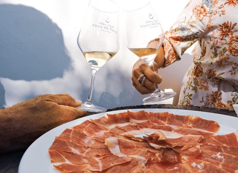 A person holding a wine glass with a floral pattern, a plate of prosciutto, and another wine glass with a similar pattern. 2