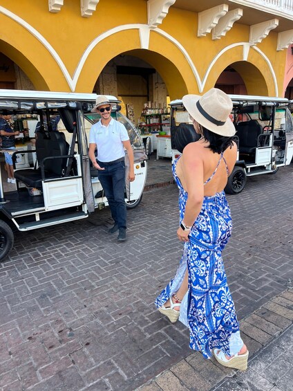 A woman in a blue patterned dress and straw hat walking past a white golf cart. 4