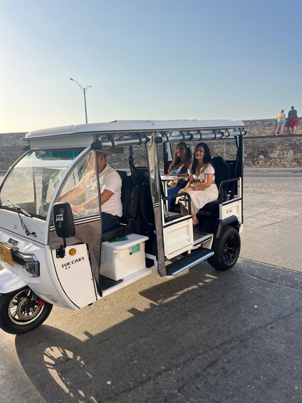 A tuk tuk with three passengers, parked on a paved area with a stone wall and a clear sky in the background. 2