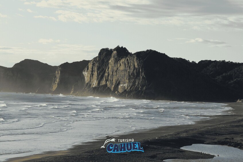 A beach with waves, a rocky cliff, and a logo for 'Turismo Cajuela.' 1
