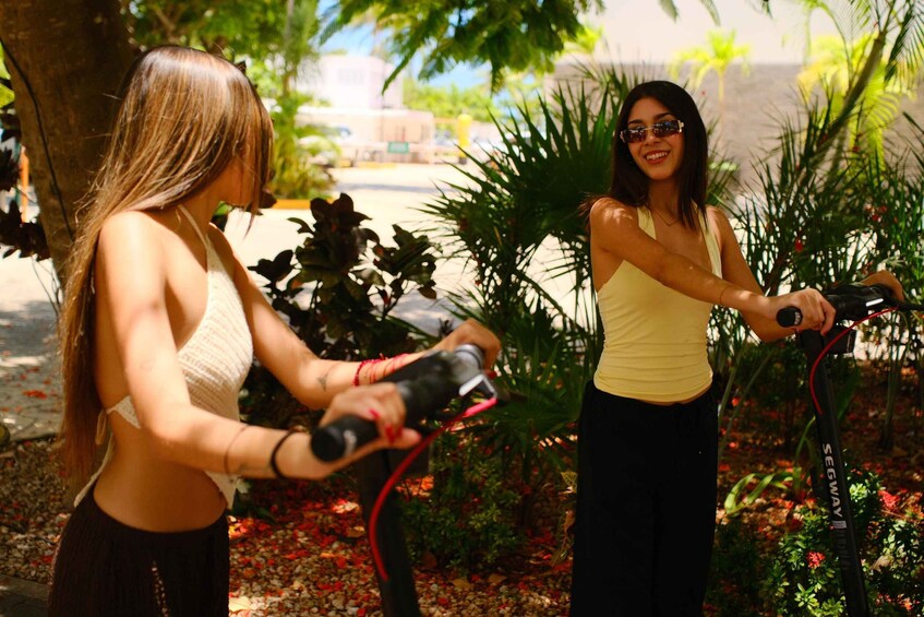 Two people on electric scooters in a garden with plants and a building in the background. 4