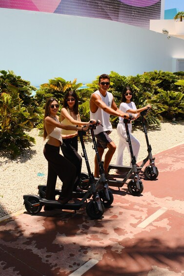 Four people riding electric scooters on a paved path with palm trees and a building in the background. 1