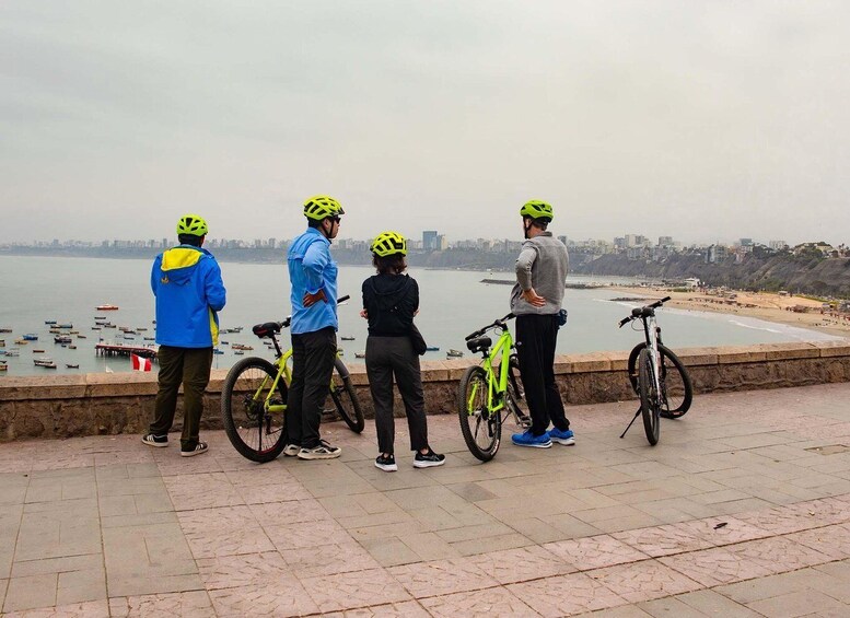 Four people standing on a waterfront promenade, wearing helmets and casual clothing, with bicycles nearby. 3
