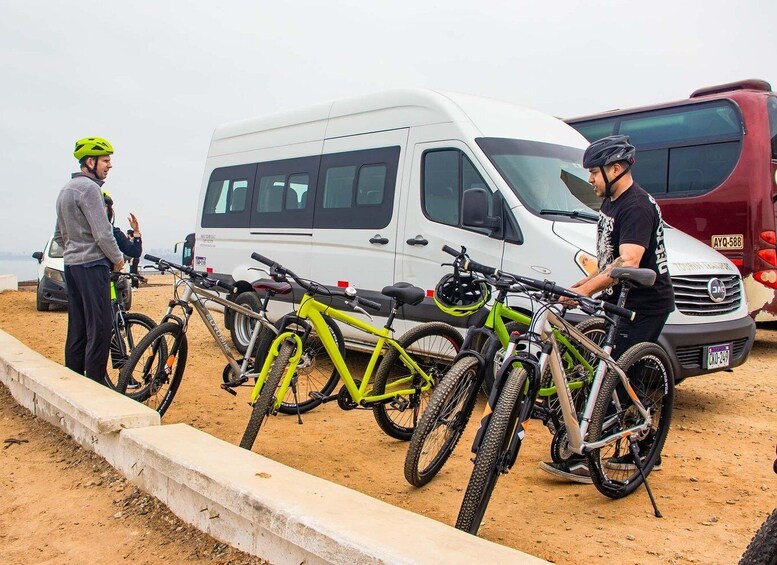 A group of people standing next to a white van and several bicycles parked on a sandy area. 2