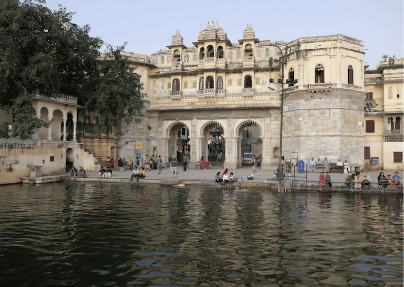A historic building with arches and domes, a water body in the foreground, and people sitting and walking around. 3