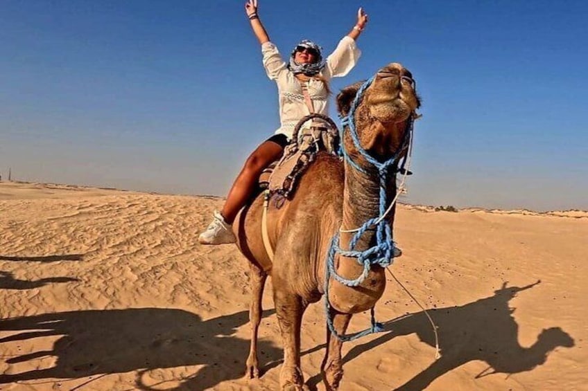 Eine Person reitet auf einem Kamel durch eine Wüstenlandschaft mit Sanddünen im Hintergrund. 1