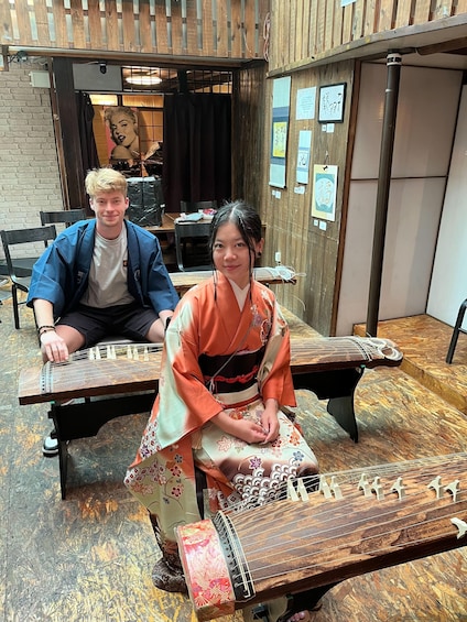 A person in traditional attire playing a stringed instrument at a wooden table in a rustic setting. 1