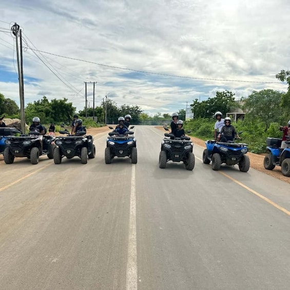A group of people riding ATVs on a paved road with trees and power lines in the background. 1