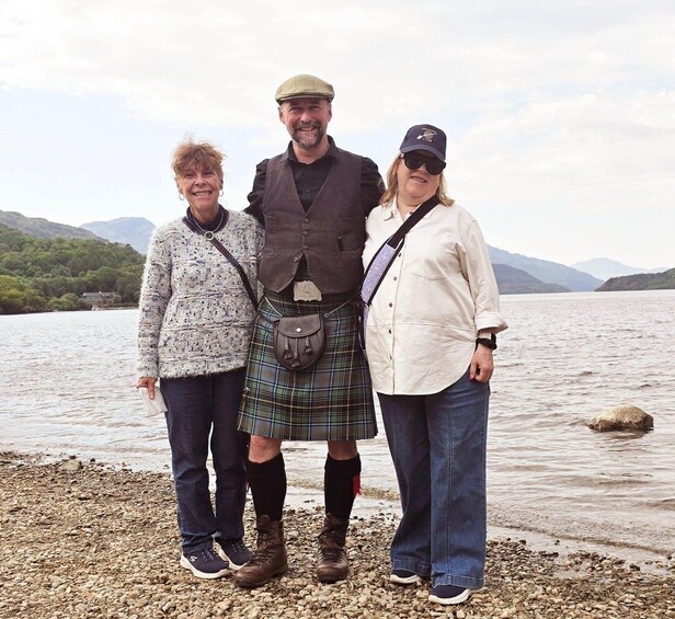 Three people standing on a rocky shore by a lake with mountains in the background. 2