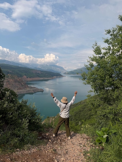 A person standing on a rocky outcrop with arms raised, overlooking a calm lake surrounded by mountains and greenery. 5