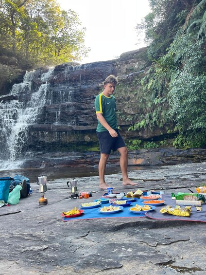 A person standing on a rocky surface near a waterfall, surrounded by a picnic setup with food and a portable stove. 3
