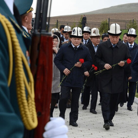A group of men in uniform and civilian attire walking with red flowers. 4