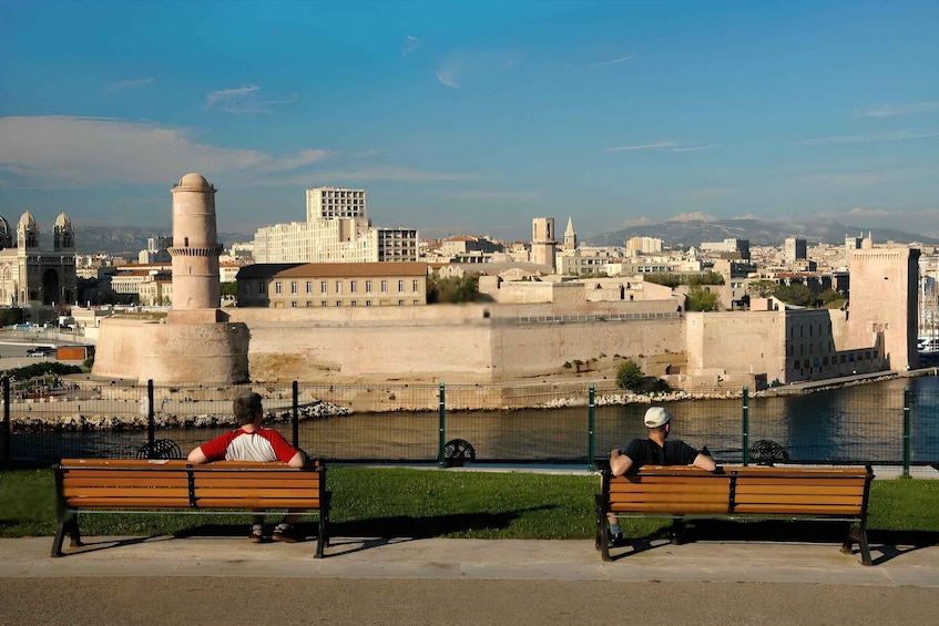 Two people sitting on wooden benches overlooking a cityscape with historical architecture and a river. 3