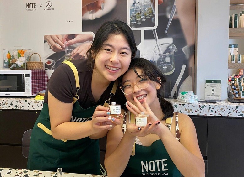 Two people in aprons holding small bottles, likely perfumes, in a store with a display of various products. 1