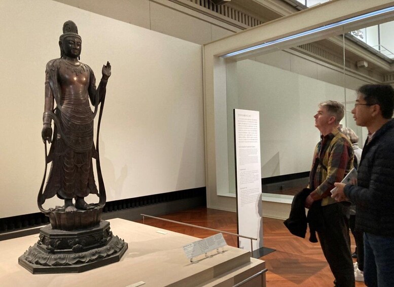 A bronze statue of a seated figure with a raised hand, displayed on a pedestal in a museum. 1