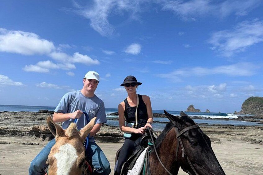 Two people riding horses on a beach with ocean and rocky formations in the background. 1