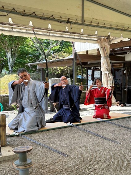 Three individuals in traditional attire, one in grey, one in blue, and one in red, seated on tatami mats under a covered area with a wooden structure in the background. 5
