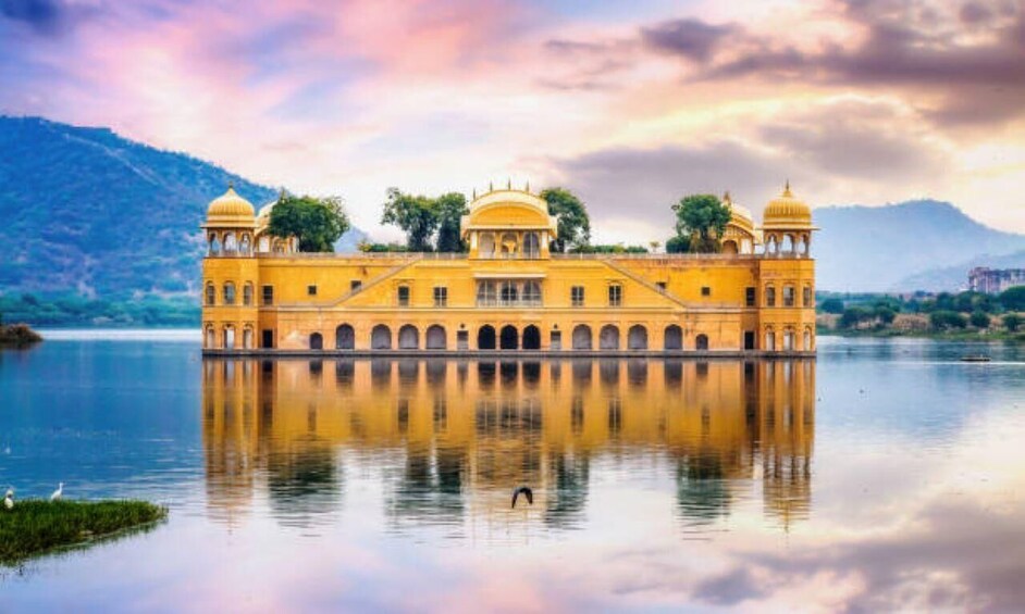 A yellow building with domes, reflecting in a calm body of water, surrounded by hills. 3
