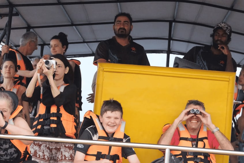 People in orange life vests, some holding cameras, on a boat with a yellow structure. 3