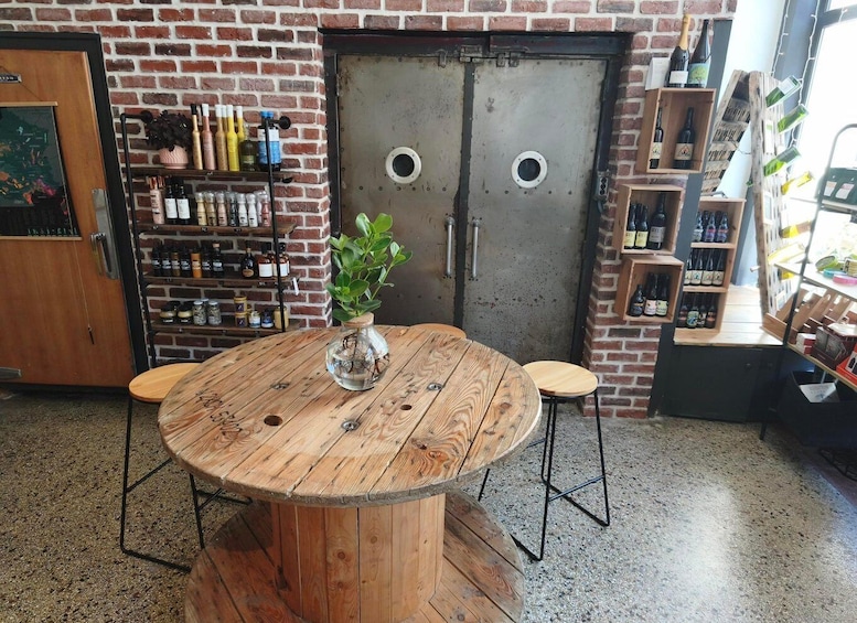 A rustic wooden table with a glass vase and plant, surrounded by shelves of bottles and jars, against a brick wall with a large metal door. 2