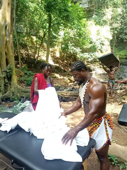 A man in traditional attire folding clothes on a table in a forested area. 1