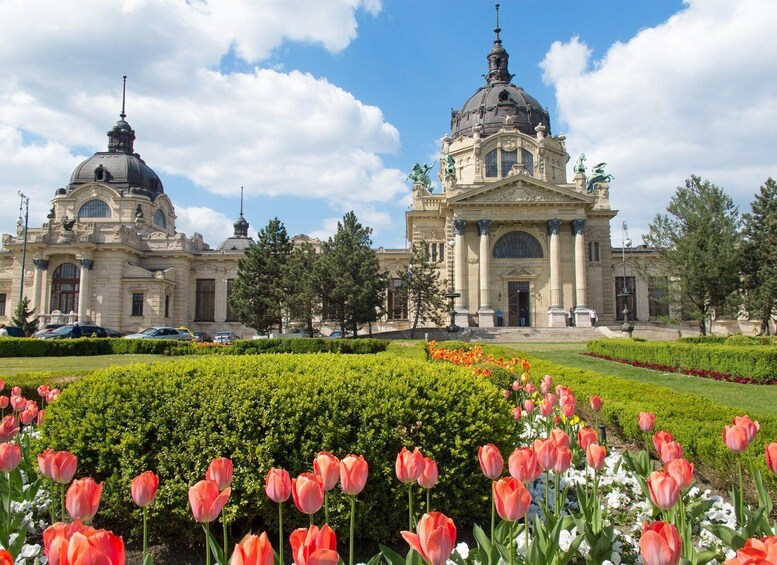 A grand building with domed roofs and statues, surrounded by a well-maintained garden with pink tulips. 2