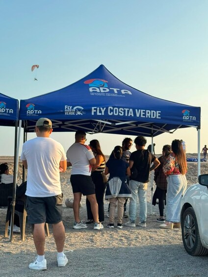 A group of people under a blue tent with 'ADTA' and 'FLY COSTA VERDE' logos, standing on a gravel surface. 2
