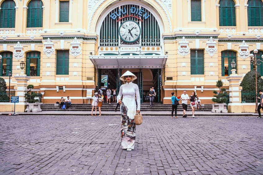 A woman in a white dress and hat walking on a cobblestone street in front of a building with a clock and the sign 'BUU DIEN.' 2