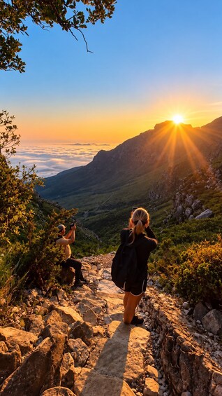 A mountainous landscape with a sunrise, rocky terrain, and two people taking photographs. 1