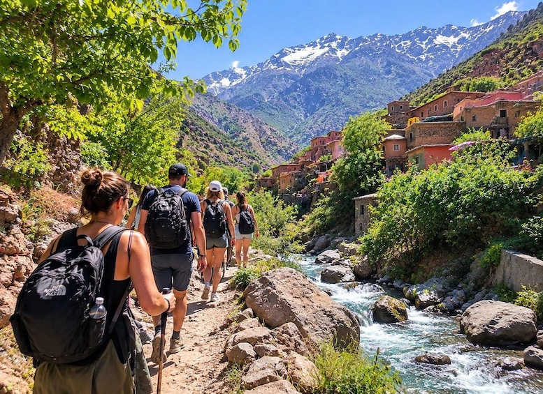 Hikers on a rocky path beside a flowing river, with colorful buildings and snow-capped mountains in the background. 1