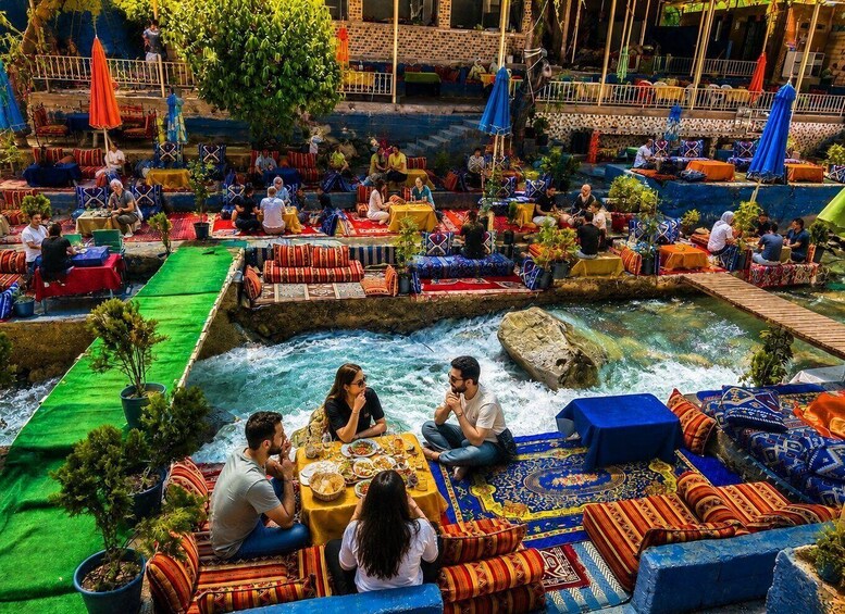 A riverside dining area with people seated at tables, surrounded by colorful cushions and potted plants. 1