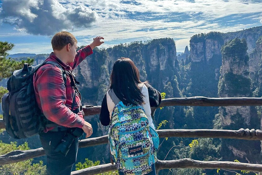 Two people standing on a wooden railing overlooking a canyon with steep cliffs and a cloudy sky. 1