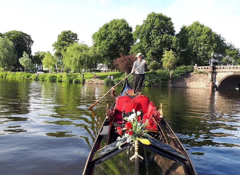 A gondola on a river with a person standing at the back, a bridge in the background, and greenery along the riverbanks. 4